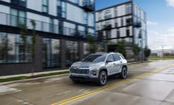 A silver SUV drives down a wet city street lined with modern glass buildings under a cloudy sky.