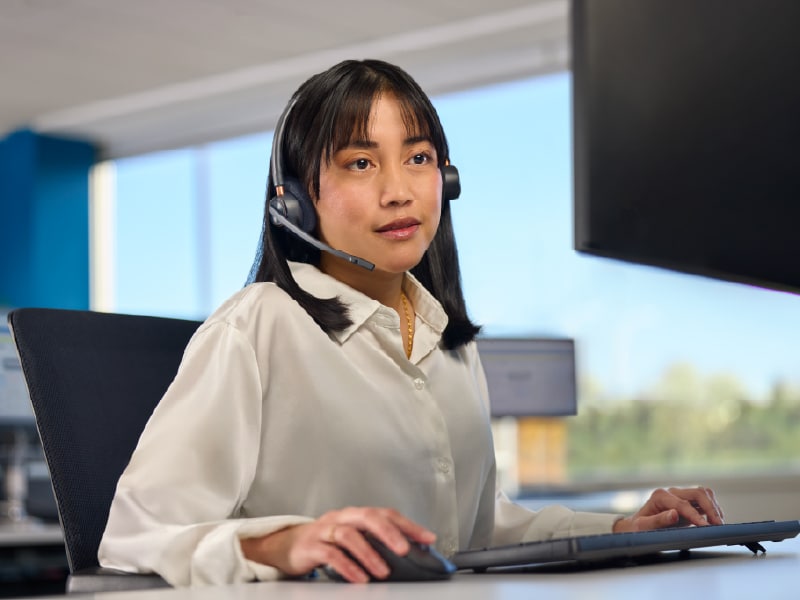 Woman in office wearing a headset, working at a computer with natural light in the background.
