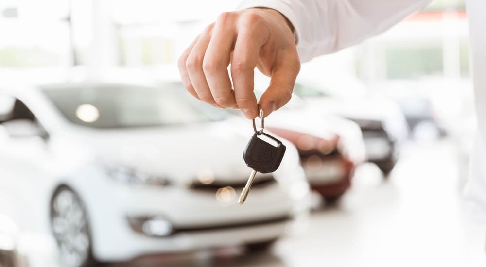 Hand holding car keys in dealership, with blurred cars in the background, symbolizing vehicle purchase.