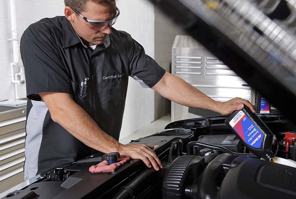 Mechanic in safety glasses adding engine oil to a car in a garage setting.