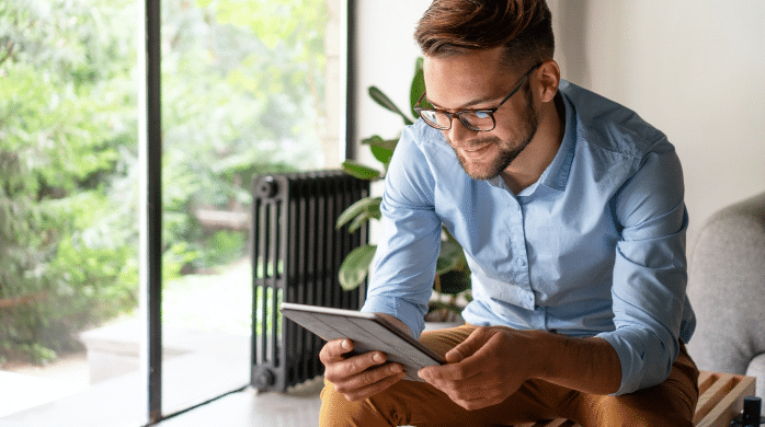 Man in glasses smiling while using a tablet in a bright room with greenery outside.