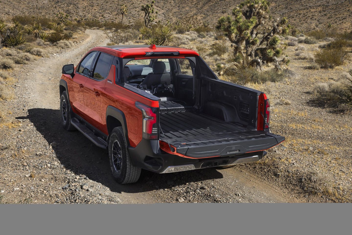 Red pickup truck with open tailgate on a rugged dirt path amid desert landscape.