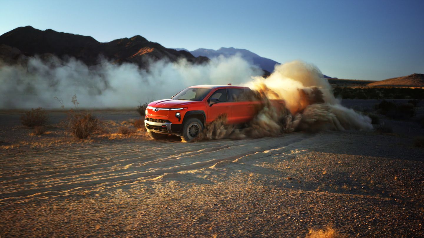 Red SUV kicks up dust driving through desert at sunset, with mountains in the background.