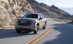 A beige GMC pickup truck drives on a curvy mountain road under a clear sky.