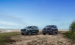 Two SUVs parked on a sandy beach under a clear blue sky, with greenery nearby.