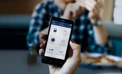 Person using a smartphone app with security icons, sitting at a table with another person in the background.