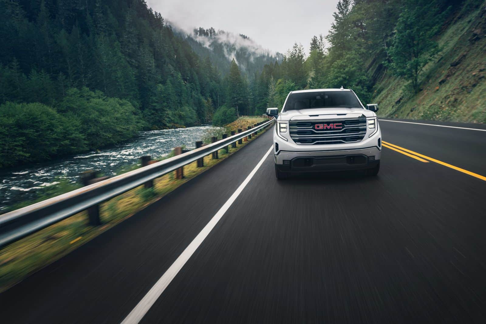 A white GMC truck drives on a scenic forest road by a river, surrounded by lush green trees.
