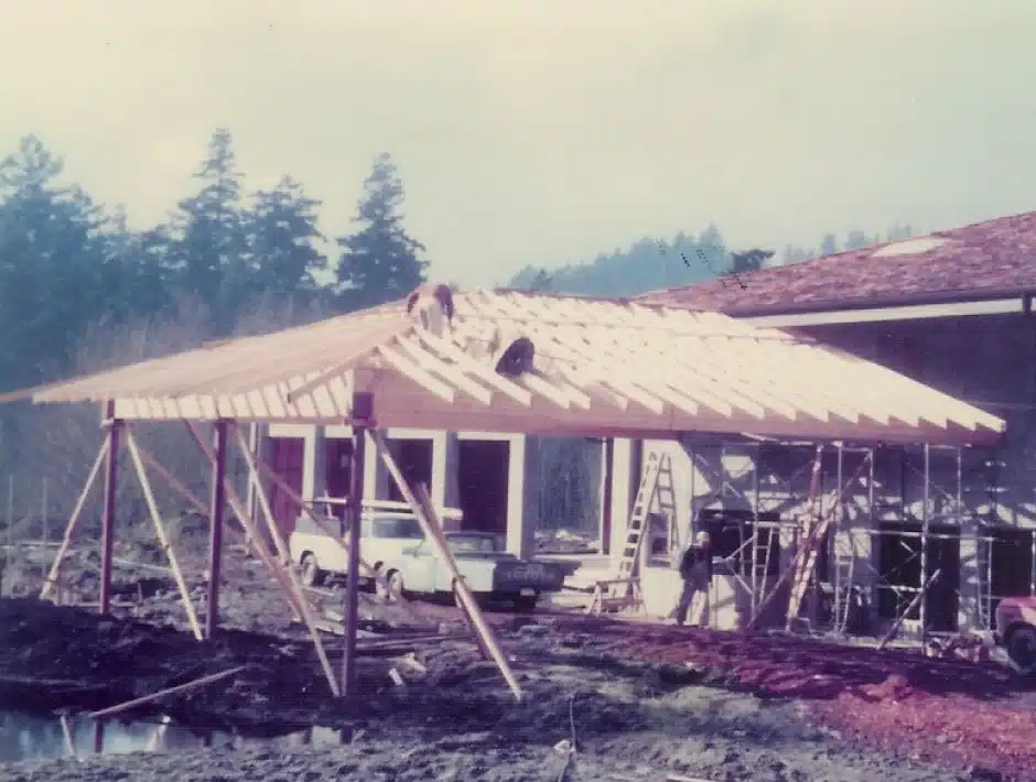 Construction site with a wooden roof frame, workers, and vehicles, surrounded by trees.