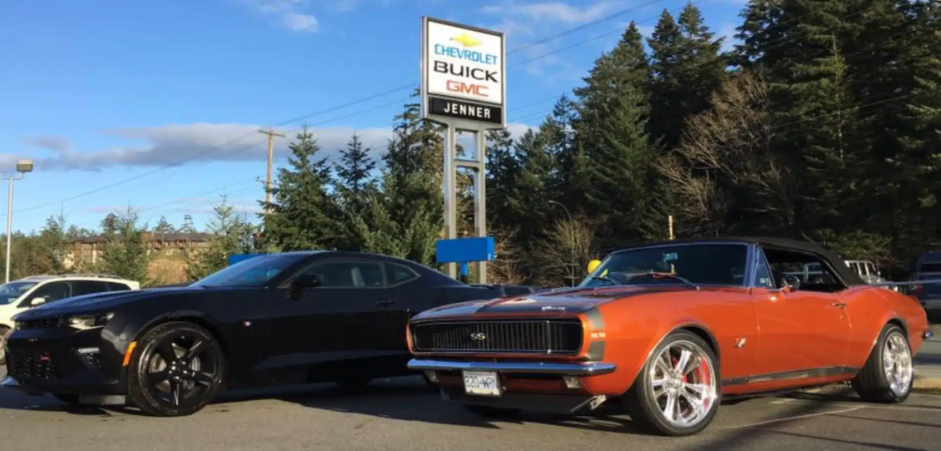 Black and orange vintage cars parked near a Chevrolet Buick GMC dealership sign, surrounded by trees.