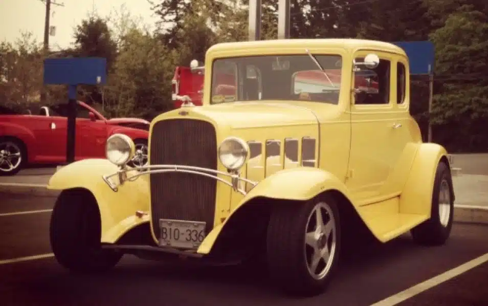Yellow vintage car parked outdoors with a visible license plate B10-336. Red convertible in the background.