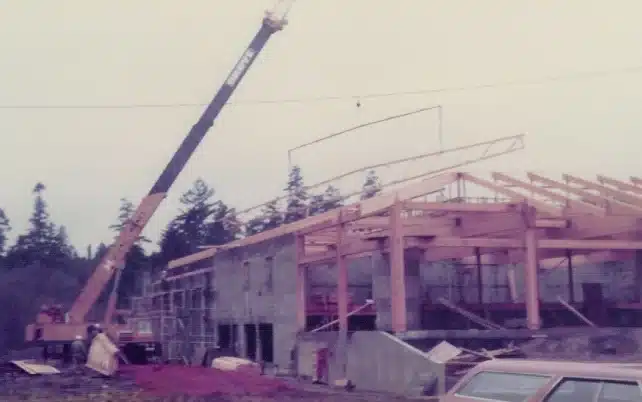 Construction site with a crane lifting a roof truss onto a partially-built structure amidst trees.
