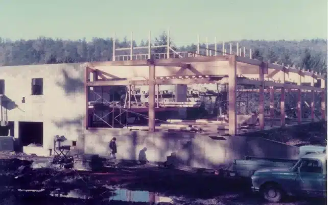 Construction site with a partially built structure, scaffolding, and parked trucks in a wooded area.